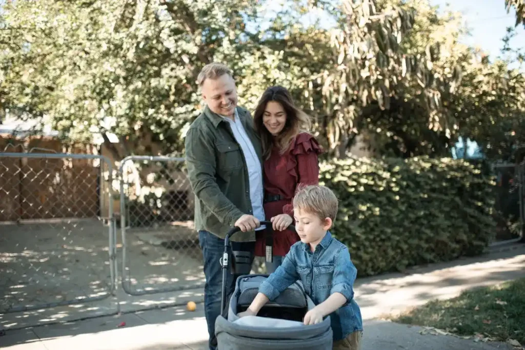 A young boy looks into a grey wagon-style stroller while his parents stand behind him, smiling.