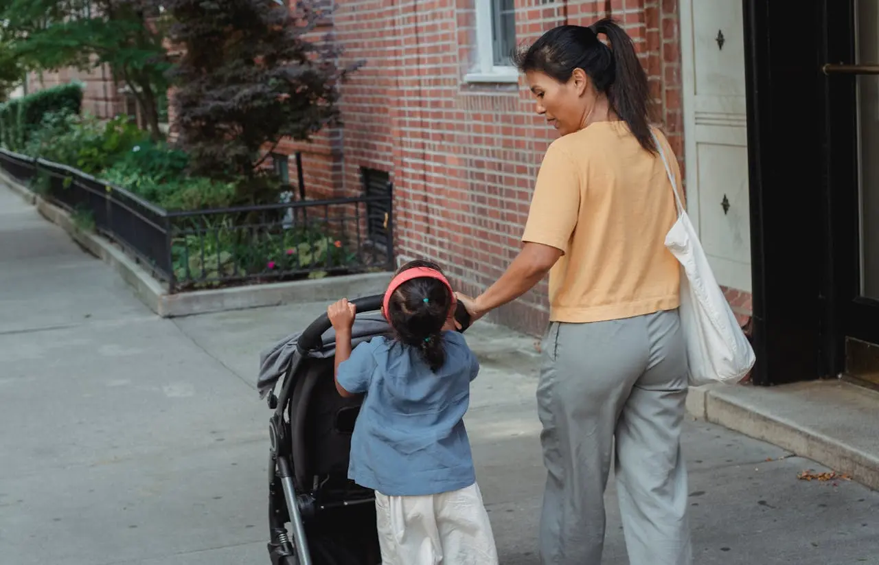 A small child stands at the handle of a double stroller while an adult guides it from behind.