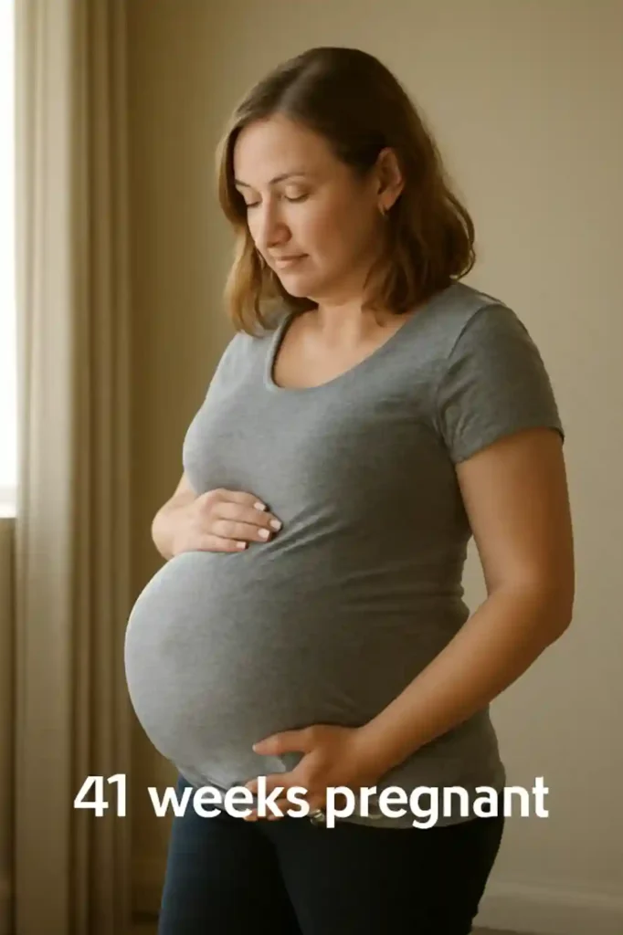 Side view of a pregnant person in a gray T-shirt standing indoors with hands supporting their belly; overlaid text reads “41 weeks pregnant.”