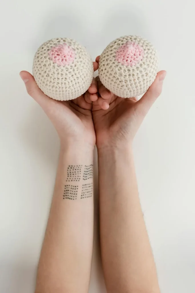 Hands holding two crocheted breast models with pink areolas, illustrating tenderness and self-care during First Trimester Sore Breasts.