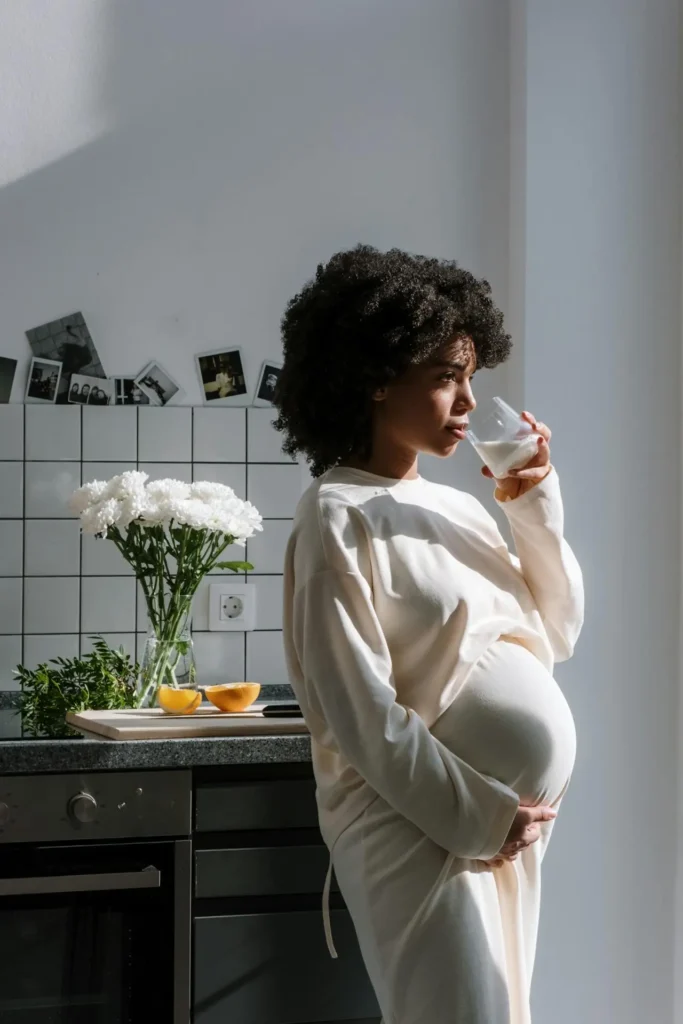 Pregnant woman in a bright kitchen sipping a glass of milk while holding her belly; flowers and sliced oranges on the counter.