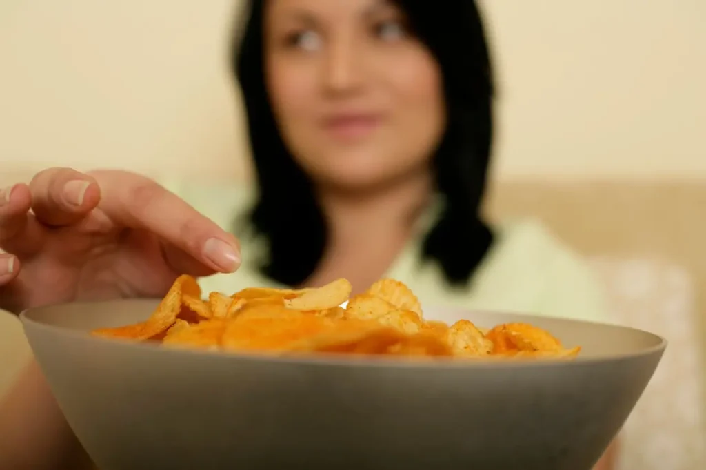 Close-up of a hand reaching into a bowl of potato chips with a blurred person in the background—an image illustrating First Trimester Food Cravings and Aversions.