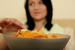 Close-up of a hand reaching into a bowl of potato chips with a blurred person in the background—an image illustrating First Trimester Food Cravings and Aversions.