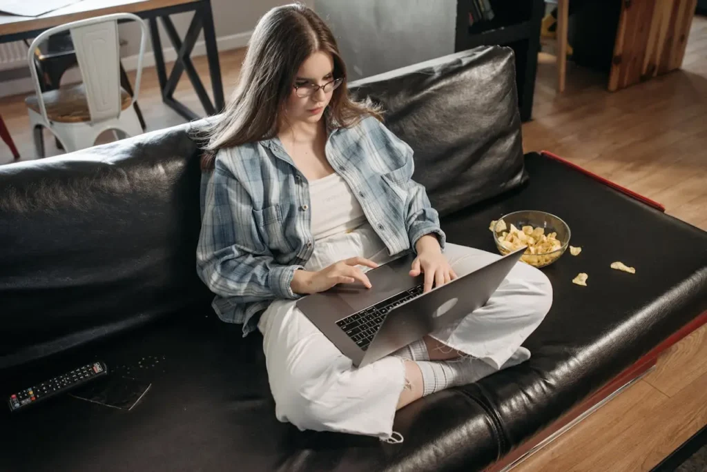 Person sitting cross-legged on a black sofa working on a laptop with a bowl of potato chips nearby—illustrating First Trimester Weight Gain, casual snacking, and rest.