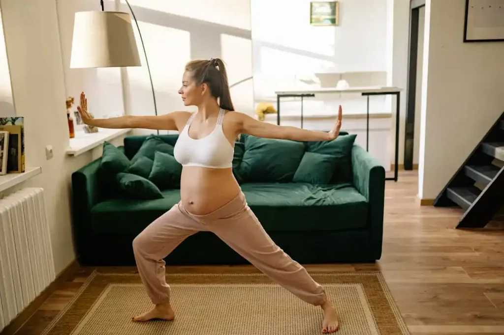 Pregnant woman in Warrior II pose at home, arms extended and knees bent, practicing breath and gentle pelvic floor engagement—Pelvic Floor Exercises for Pregnancy.