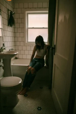 Person sitting on the edge of a bathtub in a dim bathroom, looking uncomfortable—an illustration of Second Trimester Constipation and the stress it can cause.