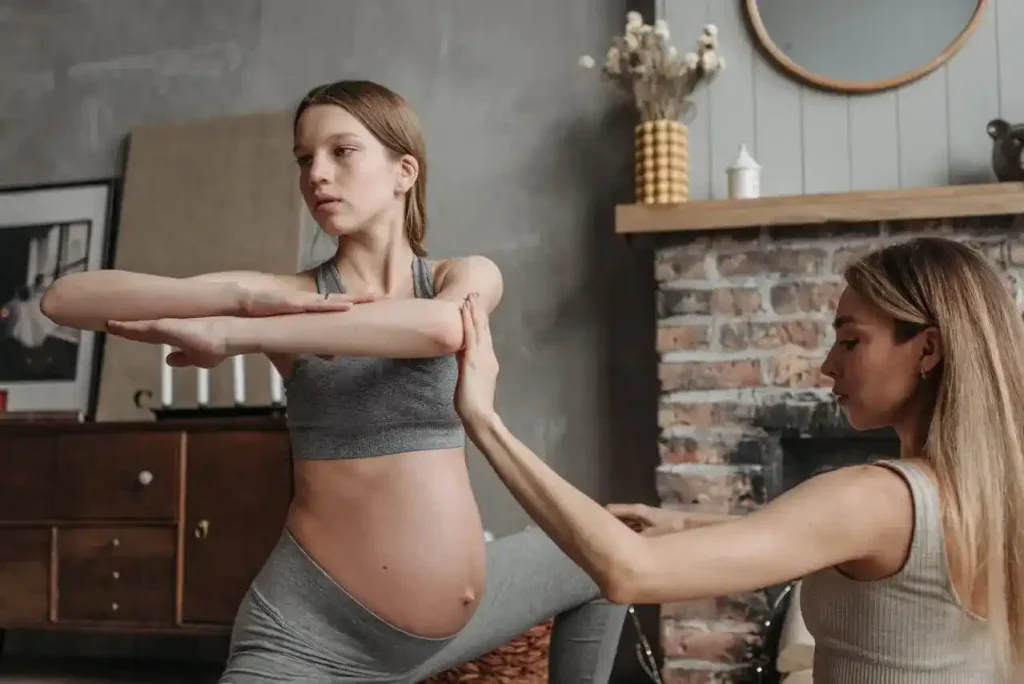 Pregnant woman in a supported lunge performing a cross-body shoulder stretch while a prenatal coach cues alignment—strength and flexibility training for pregnancy at home.