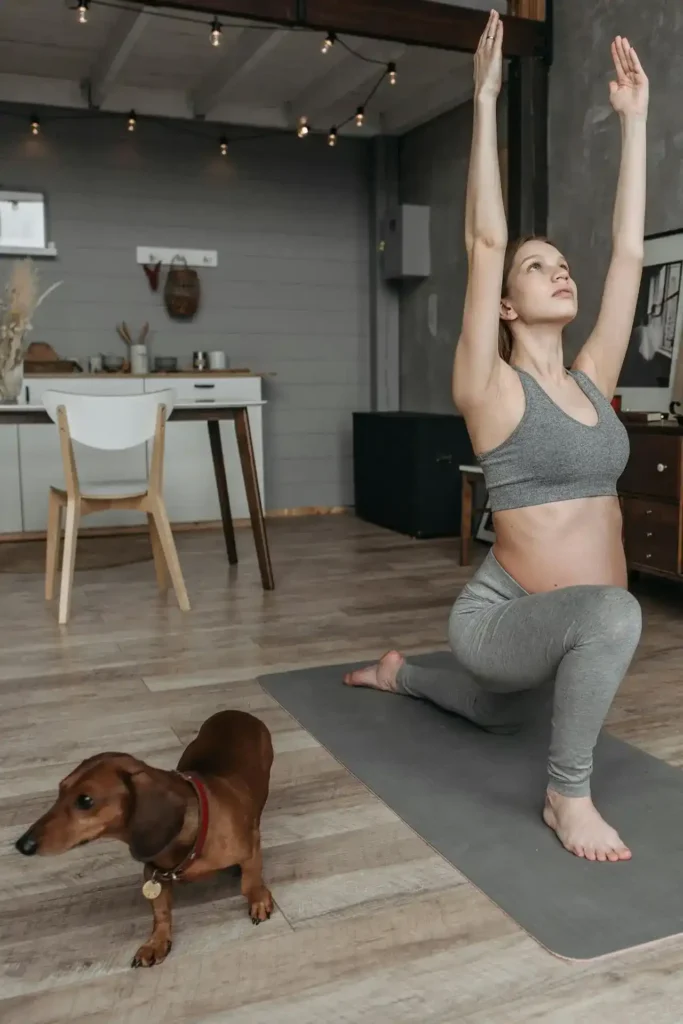 Pregnant woman in a low lunge with arms overhead on a yoga mat at home, practicing gentle hip-flexor and chest stretches for pregnancy; a small dachshund stands nearby.