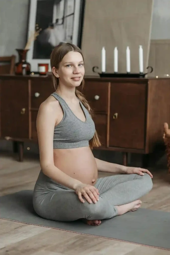 Pregnant woman sitting cross-legged on a yoga mat at home, practicing a tall-spine seated pose with relaxed breathing—Pregnancy Exercise for Pelvic and Back Pain.