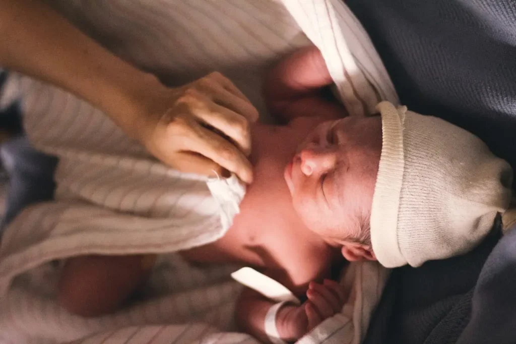 Newborn in a knit cap swaddled in hospital blankets while a caregiver adjusts the wrap—calm first minutes after an assisted delivery.