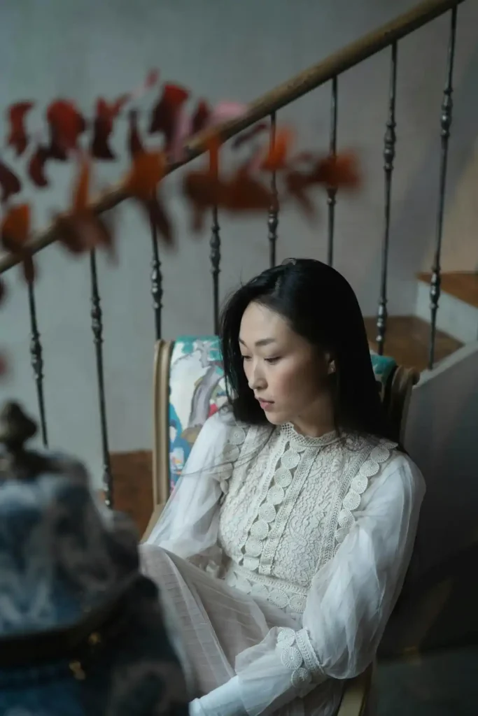 Thoughtful new mom in a lace dress sitting by a staircase, looking down in soft natural light—visualizing the emotions behind baby blues vs postpartum depression.