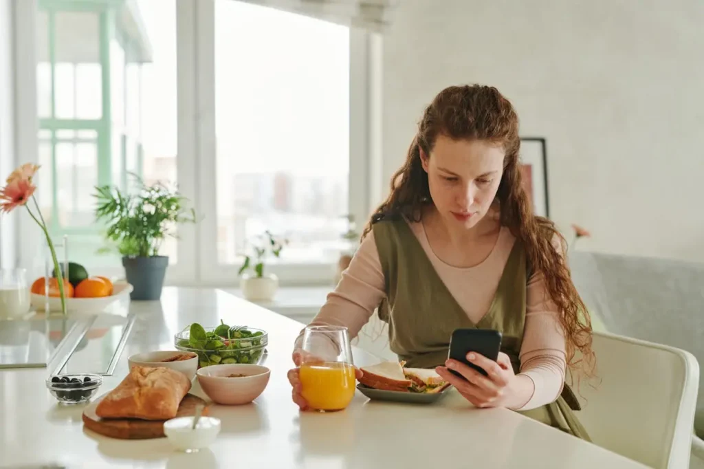 Expectant mom at a bright kitchen table eating a balanced breakfast—whole-grain sandwich, leafy green salad, yogurt, berries, and orange juice—while checking her phone.