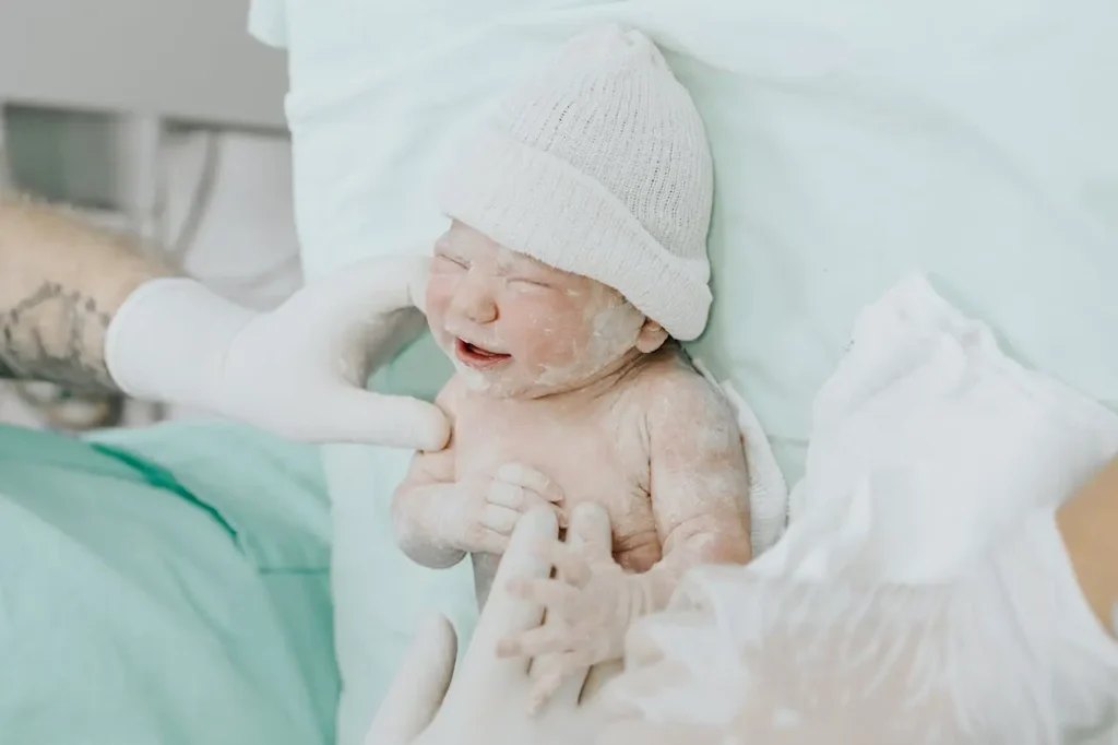 Newborn covered in vernix, wearing a knit cap, held by gloved clinicians on the operating table just moments after a C-section
