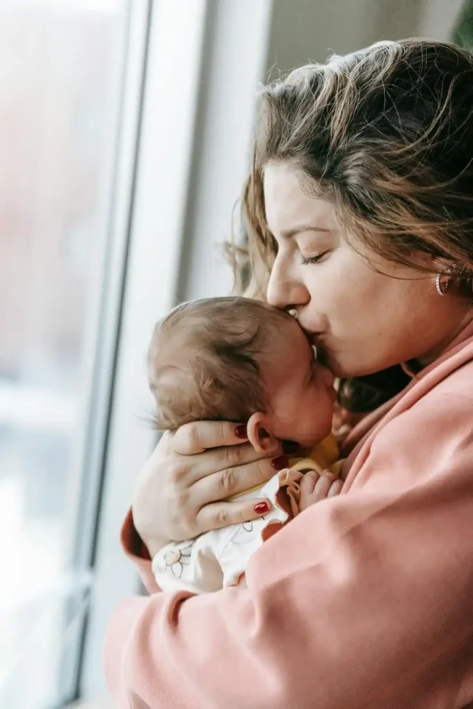 New mom in a pink sweater kisses her newborn by a bright window—quiet bonding during C-section recovery.