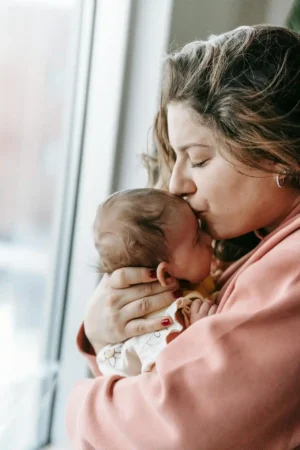 New mom in a pink sweater kisses her newborn by a bright window—quiet bonding during C-section recovery.