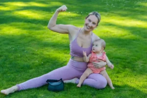 Smiling new mom in a lavender workout set sits on the grass with her baby, flexing her arm—celebrating easing back into exercise after delivery.