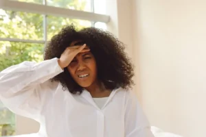 Person in a bright room holding a hand to their forehead with a pained expression, illustrating First Trimester Morning Sickness and queasy fatigue.