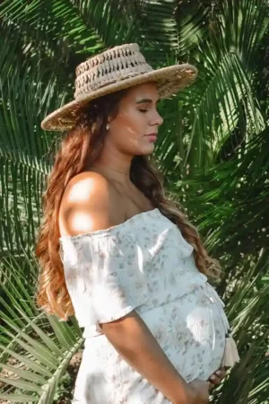 Pregnant person in a straw hat standing among palm leaves with soft sunlight on her face, illustrating First Trimester Skin Changes such as pigmentation and sensitivity.
