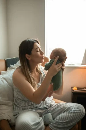 New mom in striped pajamas lifts and kisses her newborn by a bright window—quiet bonding while recovering and monitoring Postpartum bleeding (lochia).