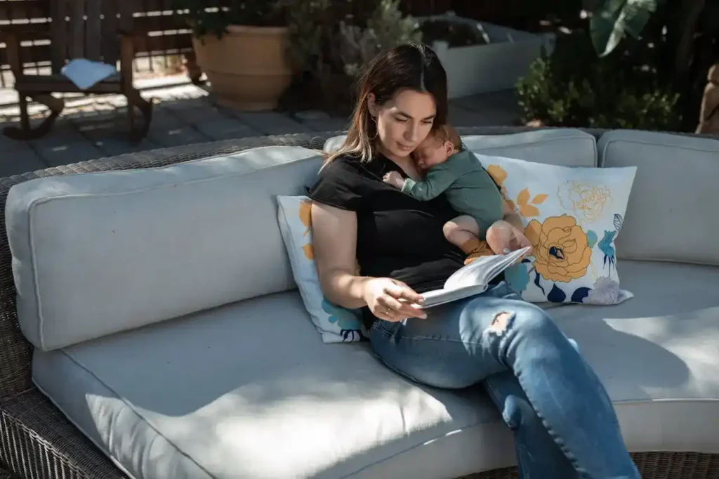 New mom on patio sofa, newborn on chest, reading during recovery from postpartum Vaginal soreness and tearing.
