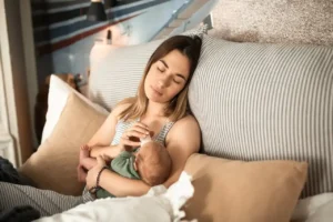 Tired new mom reclines on pillows, bottle-feeding her newborn—a calm reminder that rest and nourishment are part of healthy Postpartum Weight Loss.