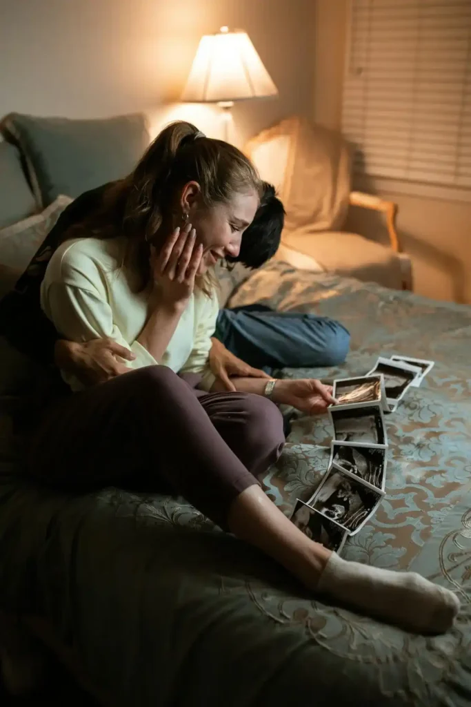 Couple sitting on a bed, smiling and emotional as they look at a strip of ultrasound photos under warm lamp light.