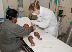 Clinician in protective gear examines an infant on a hospital bed while a caregiver comforts them—clinic visit related to Baby Common Illnesses.