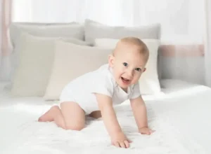 Smiling baby in a white onesie on hands and knees on a bed, showing Baby Crawling posture in a bright, cozy room.