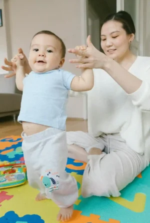 Smiling baby practicing standing on a colorful play mat while a parent holds their hands for support, illustrating Baby Development Milestones by Month.