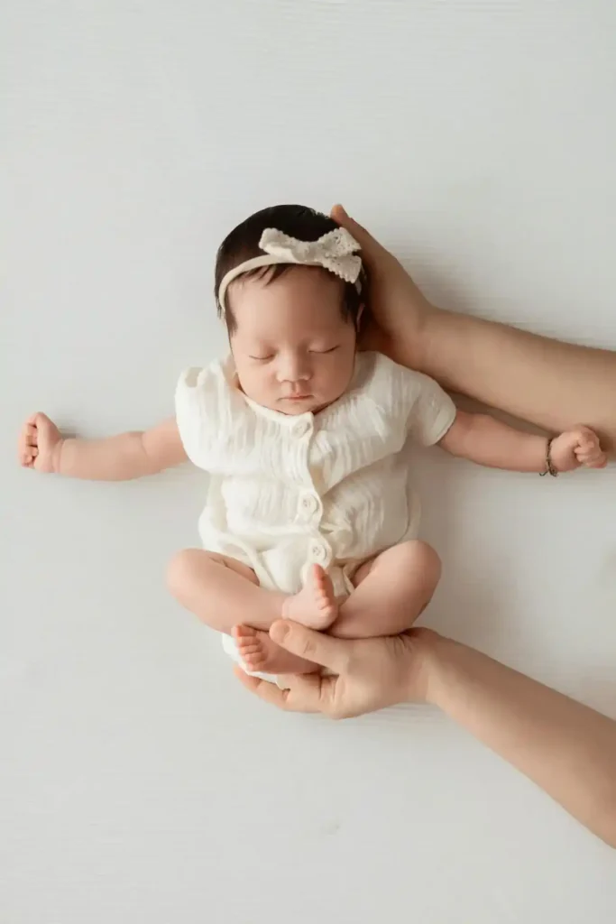 Newborn in a white onesie and headband lying on their back while a caregiver supports the head and feet—a calm moment linked to Baby Vaccination day.
