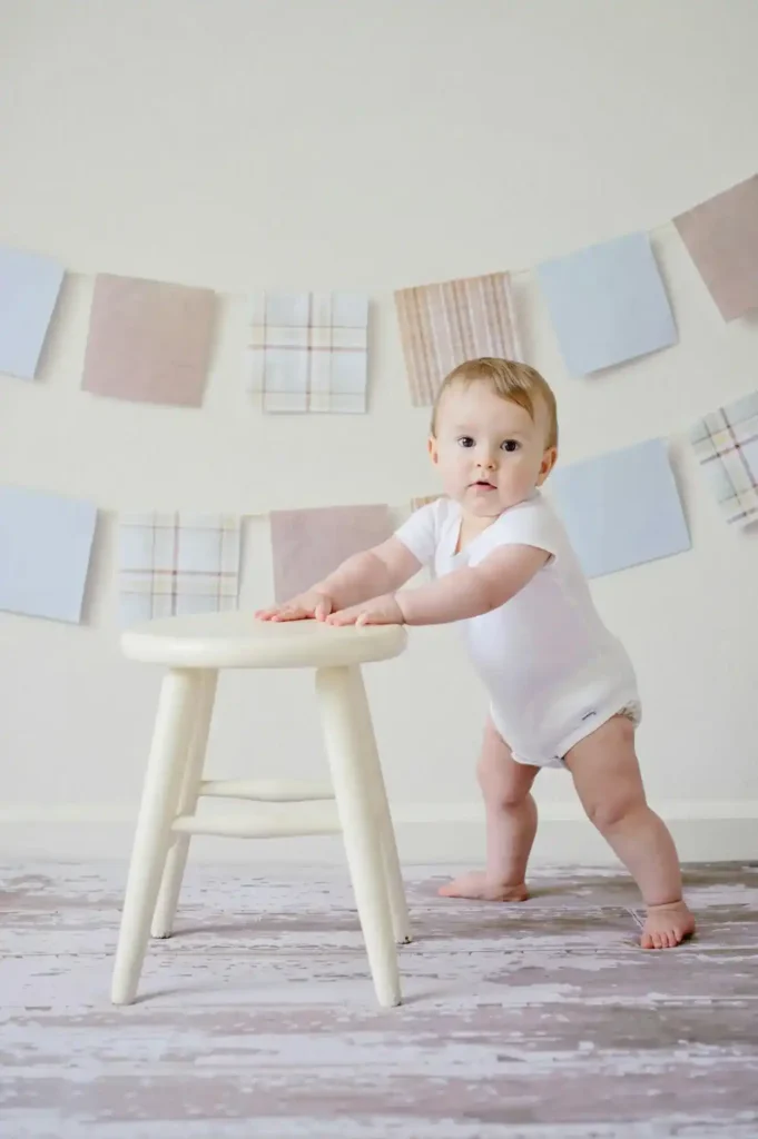 Baby in a white onesie standing and holding a small stool—Baby Walking practice at the cruising stage in a bright playroom.