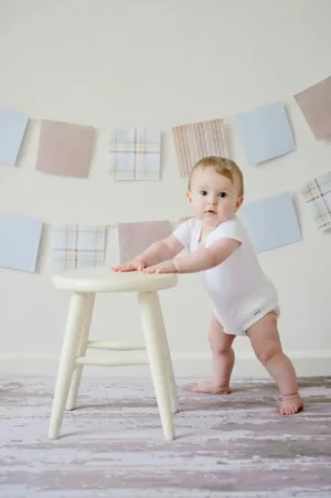 Baby in a white onesie standing and holding a small stool—Baby Walking practice at the cruising stage in a bright playroom.