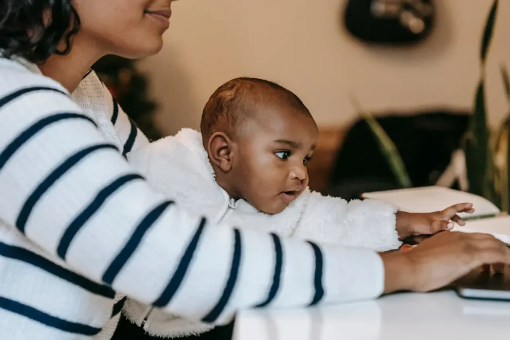A close-up of a mother working on a laptop at a white desk while holding her baby in her lap. The mother wears a striped sweater, and the baby, wearing a fluffy white jacket, looks attentively toward the screen or task, illustrating the challenge of balancing work childcare and home life.