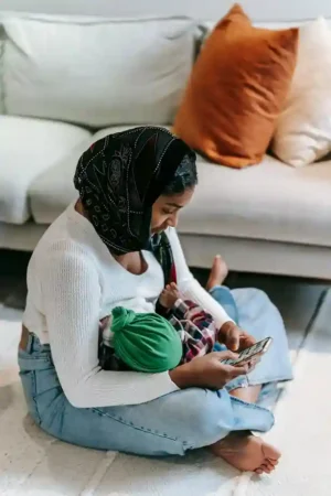 Mom sitting cross-legged on a rug, practicing Breastfeeding for Baby in a cozy living room, gently cradling her infant while glancing at her phone.