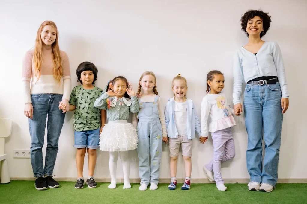 Two female teachers or caregivers, one with long red hair and the other with short curly dark hair, stand on opposite ends of a row of five diverse young children. Everyone is smiling and holding hands, posing against a plain white wall above a green floor. The scene represents a positive school or daycare environment.
