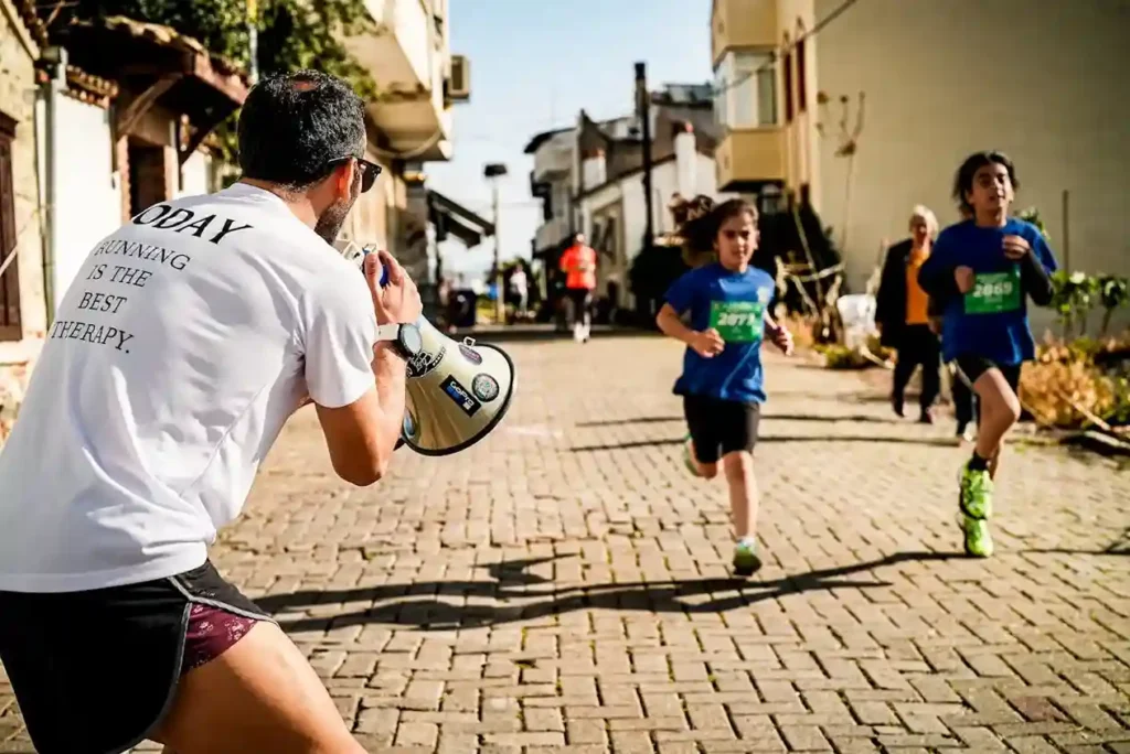 A man wearing a white shirt that reads "TODAY RUNNING IS THE BEST THERAPY" is enthusiastically shouting encouragement through a megaphone toward two children who are running in a race on a cobblestone street. The children are wearing blue race shirts with numbers.