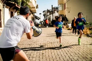A man wearing a white shirt that reads "TODAY RUNNING IS THE BEST THERAPY" is enthusiastically shouting encouragement through a megaphone toward two children who are running in a race on a cobblestone street. The children are wearing blue race shirts with numbers.