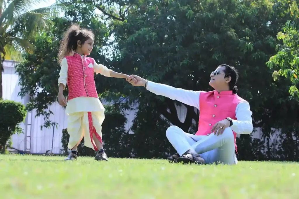 A man sitting cross-legged on a sunny green lawn holding the hand of a young child who is standing. They are both wearing matching traditional Indian-style outfits featuring pink vests over white and cream clothing. The image captures a loving parent-child connection outdoors, suggesting a strong bond that informs finding your parenting style.