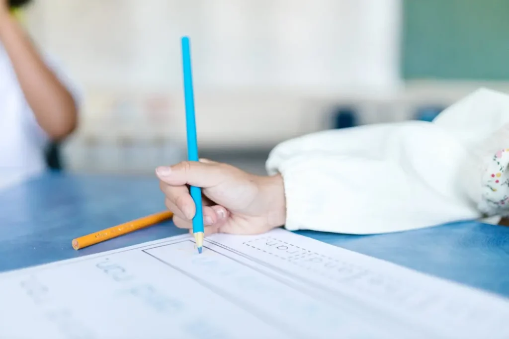 A close-up shot of a young child's hand holding a blue pencil with a correct tripod grip, tracing letters on a handwriting practice worksheet on a blue table in a classroom setting. Another orange pencil rests beside the paper.