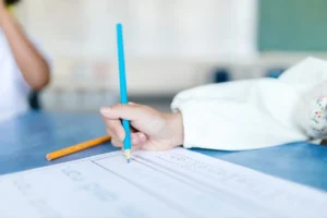 A close-up shot of a young child's hand holding a blue pencil with a correct tripod grip, tracing letters on a handwriting practice worksheet on a blue table in a classroom setting. Another orange pencil rests beside the paper.
