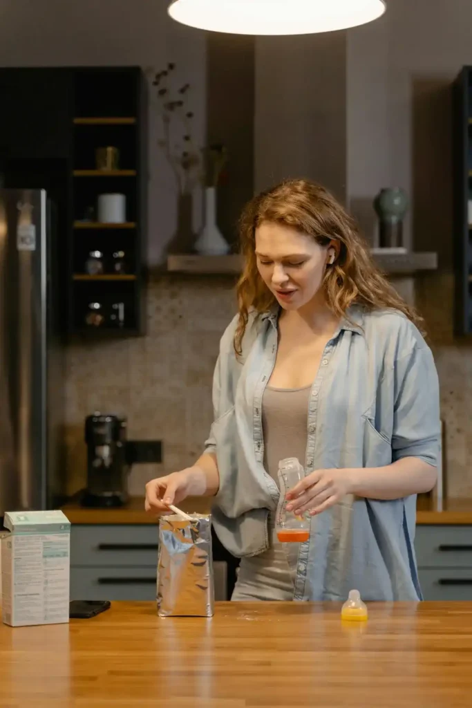 Parent measuring powdered formula into a baby bottle in a home kitchen—Formula Feeding preparation with scoop, box, and nipple on the counter.