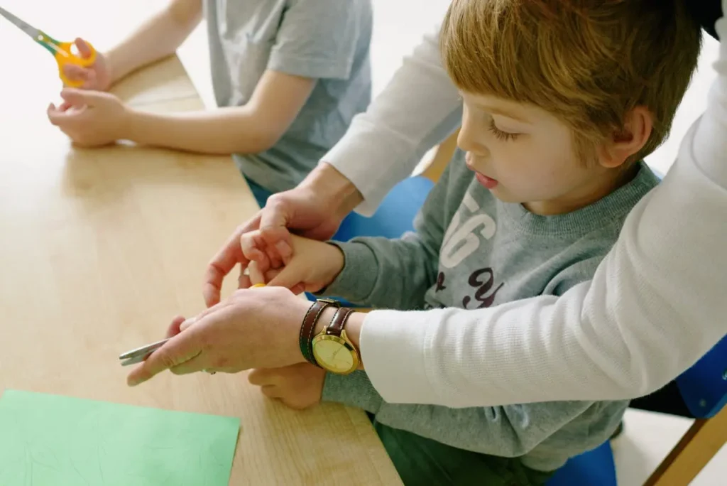 A close-up shot of an adult helping a young, light-haired boy use scissors to cut green paper. The adult's hands gently guide the child's hands and the small scissors, indicating instruction or assistance with fine motor coordination.