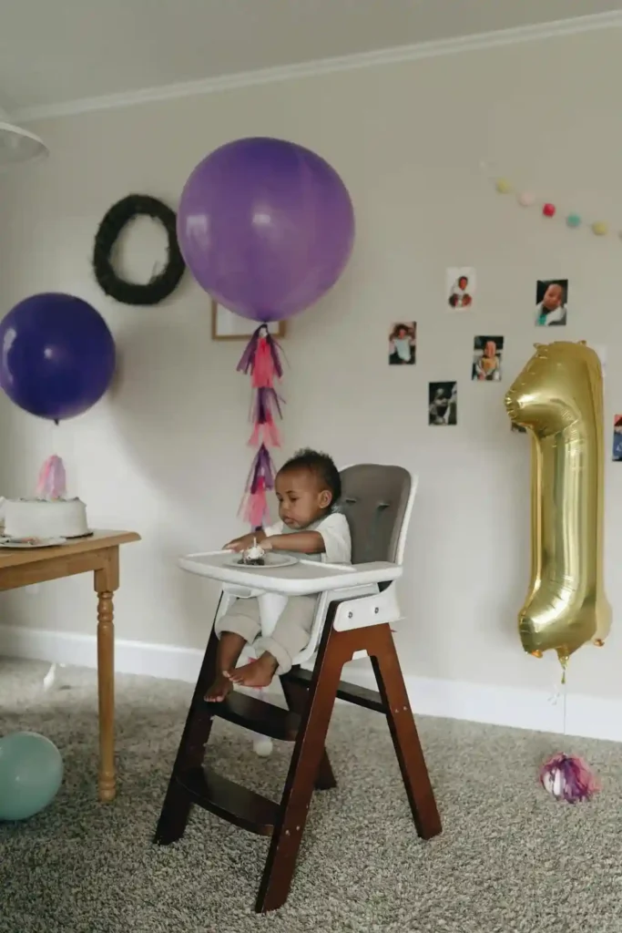 A baby, likely celebrating their first birthday, is seated in a modern wooden high chair, looking down at a small treat. The background is decorated with large purple balloons, a party banner, small framed photos on the wall, and a large gold "1" balloon, alluding to their age and developmental stage.