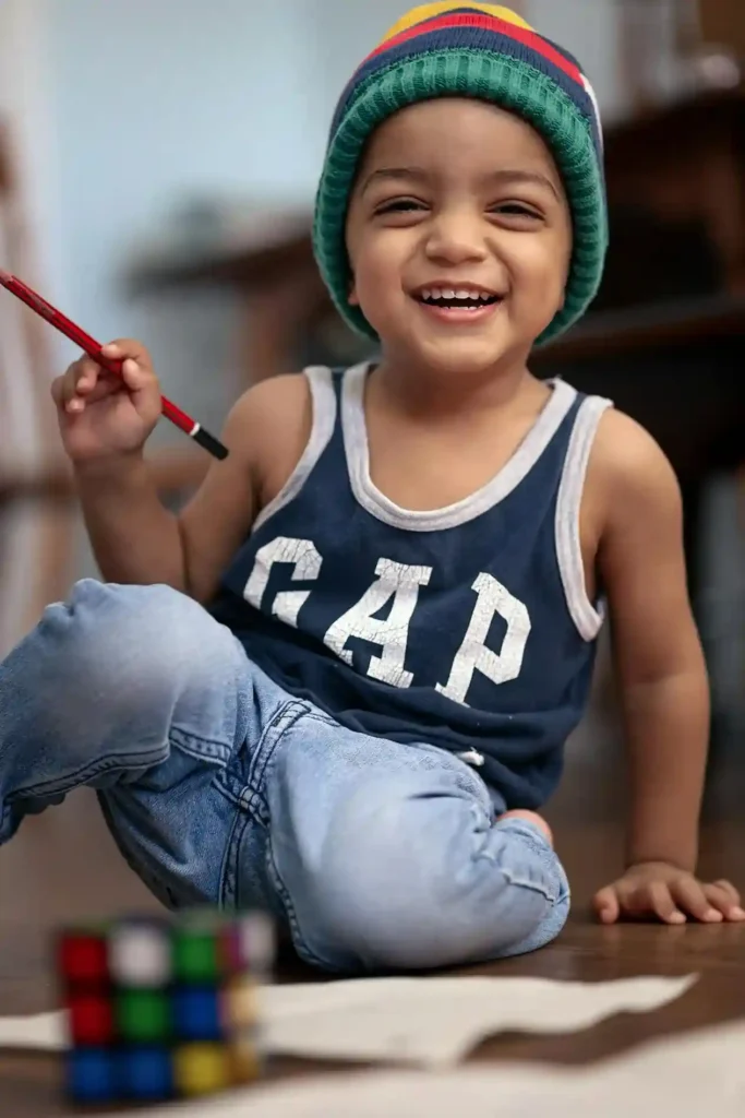 A brightly smiling young toddler boy, wearing a striped beanie and a dark blue tank top, is seated cross-legged on a wooden floor, happily holding a red pencil or crayon up in his hand. A blurred sheet of paper and a small cube puzzle are visible in the foreground.