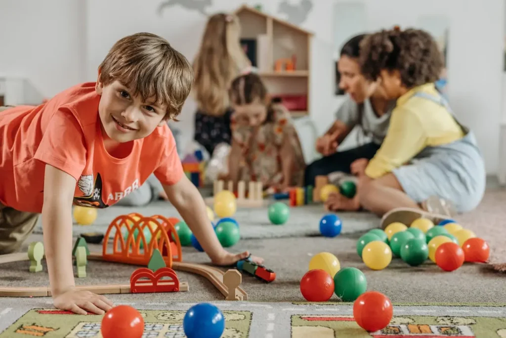 A young child playing with colorful toys in a daycare classroom while other children and a caregiver interact in the background.