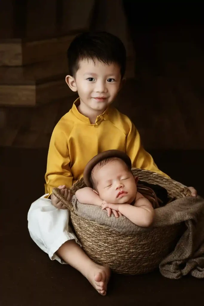 A young boy, who appears to be an older sibling, is sitting on the floor in traditional clothing and smiling proudly next to a newborn baby peacefully sleeping in a rustic woven basket. The image symbolizes a big family change like welcoming a new sibling.