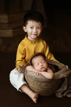 A young boy, who appears to be an older sibling, is sitting on the floor in traditional clothing and smiling proudly next to a newborn baby peacefully sleeping in a rustic woven basket. The image symbolizes a big family change like welcoming a new sibling.