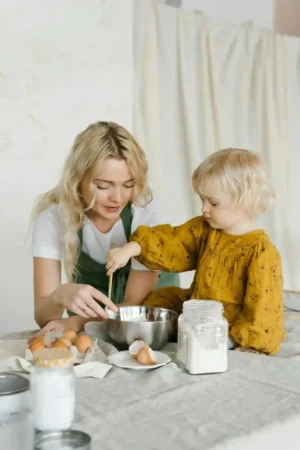 A mother wearing a green apron helps her young, blonde child in a yellow dress stir ingredients in a stainless steel mixing bowl on a kitchen table. Ingredients like eggs and flour are visible, representing a shared baking activity.