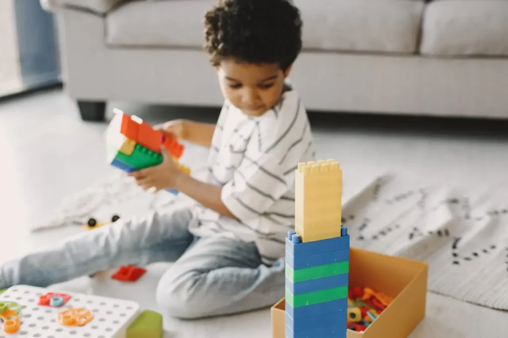 A young boy with curly dark hair is sitting on the floor in a bright living room, engaged in building a structure using large colorful plastic building blocks. A tall, partially constructed tower stands prominently in the foreground.