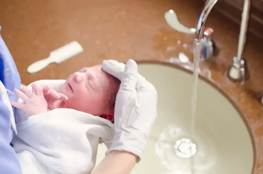Healthcare professional gently washing a newborn’s head under warm running water during a safe newborn bathing routine.